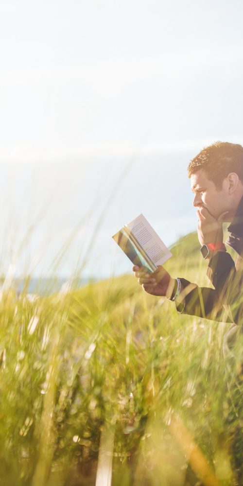 man reading book on beach near lake during daytime